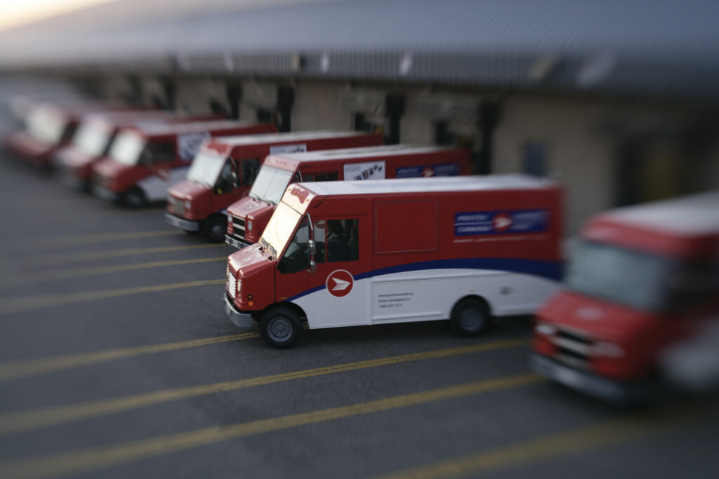 Demi-douzaine de camions « step-vans » de Postes Canada dans un stationnement, dont un qui quitte.