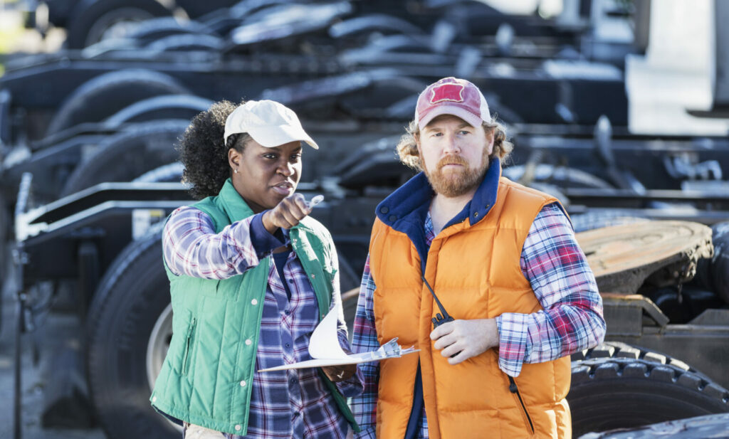 Femme noire et homme blanc aux cheveux roux dans la cour d’une entreprise de camionnage avec camions bob-tail en arrière-plan.