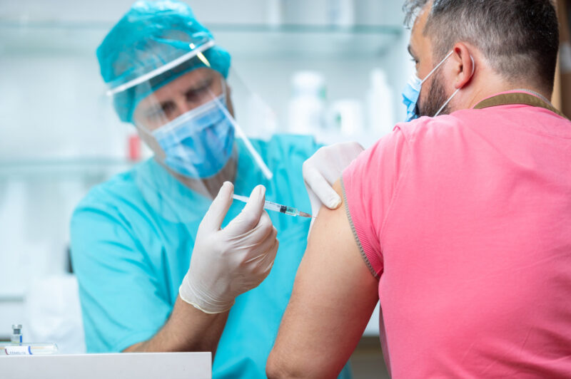 Un homme vu de dos se fait vacciner le bras gauche par un homme portant masque et visière.