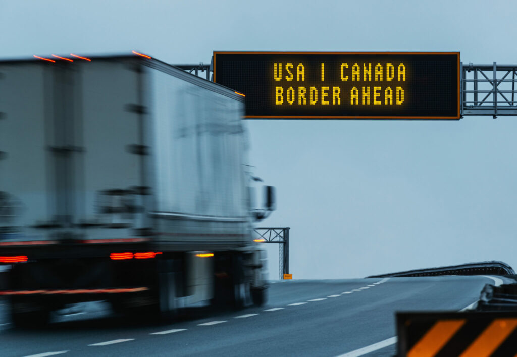Camion porteur vu de l’arrière passe sous un panneau qui indique qu’il approche de la frontière canadienne.