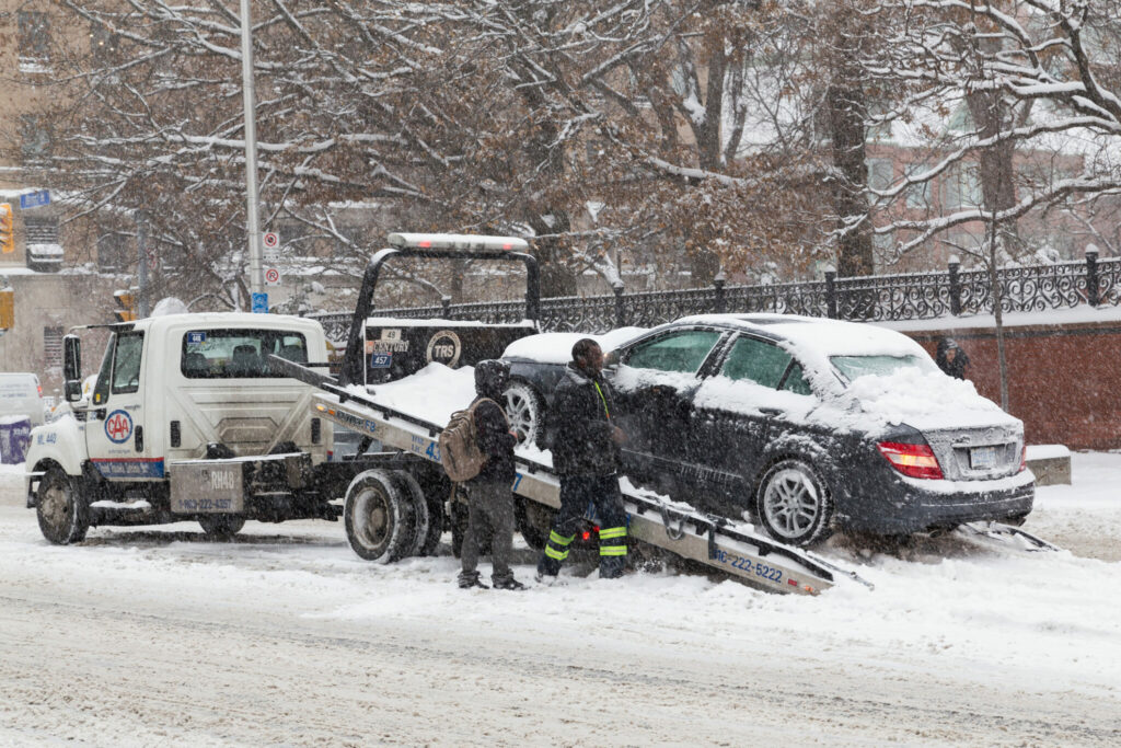 Dans une tempête de neige, deux hommes à côté d’une remorqueuse où se trouve une voiture